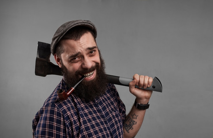 Foto grátis homem feliz com o cachimbo na boca e o machado na mão. sorridente cara atraente com barba em fundo cinza no estúdio. satisfação do conceito de vida.