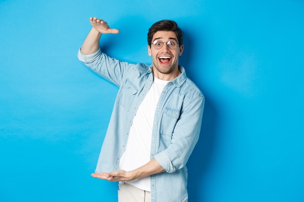 Homem feliz de óculos, mostrando um objeto de tamanho grande, dando forma a uma caixa grande, em pé sobre um fundo azul e sorrindo