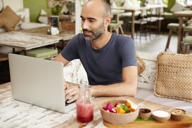 Homem feliz, digitando a mensagem no laptop enquanto toma um smoothie fresco no restaurante na calçada.