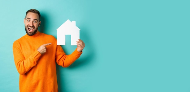 Foto grátis homem feliz do conceito imobiliário apontando o dedo para a casa de papel e sorrindo em pé sobre a luz bl