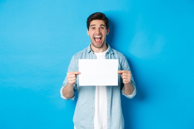 Homem feliz e atraente mostrando um pedaço de papel para sua placa de logotipo, surpreso contra um fundo azul