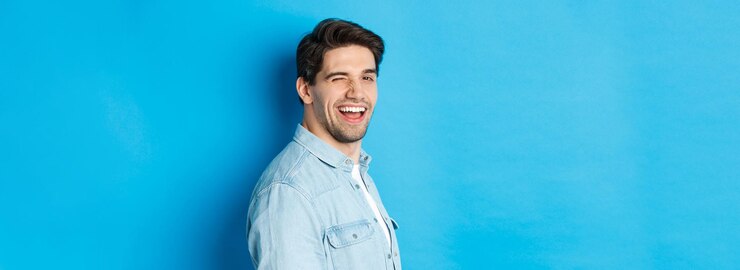 Foto grátis homem feliz e confiante vira a cabeça para a câmera piscando e sorrindo em pé sobre fundo azul