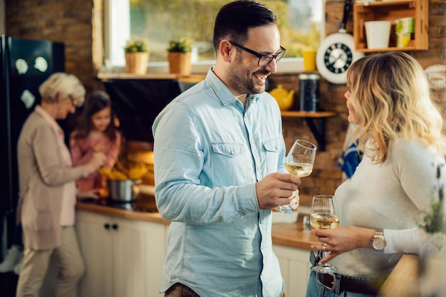 Homem feliz se comunicando com sua esposa enquanto bebe vinho na cozinha Sua filha e avó estão preparando comida na cozinha