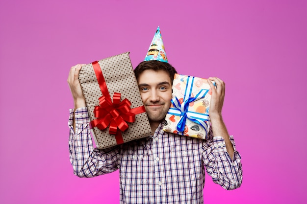 Homem feliz segurando presentes de aniversário em caixas sobre parede roxa.