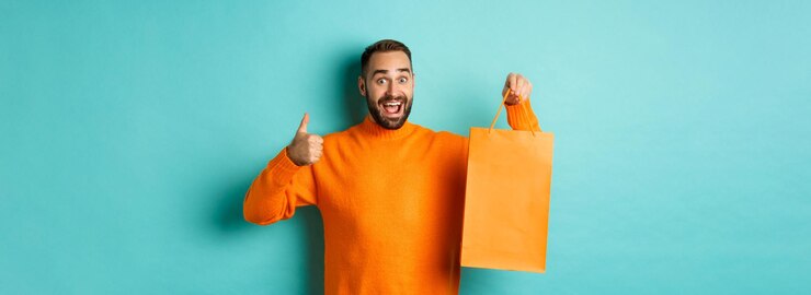 Foto grátis homem feliz segurando sacola de compras laranja e regozijando-se com desconto e comemorando mostrando polegares para cima