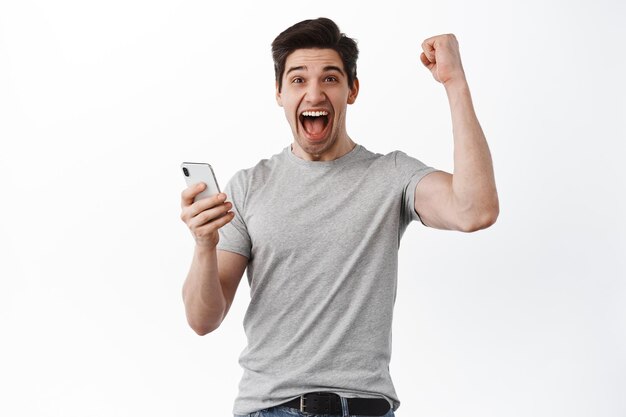Homem feliz segurando smartphone e comemorando, ganhando prêmio online, grita sim e regozijando-se, triunfando do sucesso, parece satisfeito, fundo branco