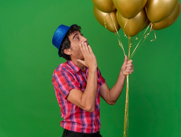 Foto grátis homem festeiro de meia-idade impressionado em vista de perfil segurando balões, olhando para eles e sussurrando isolados na parede verde