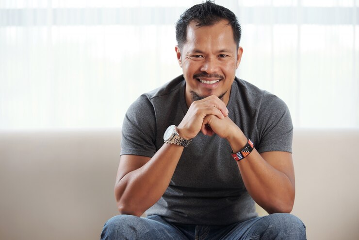 Foto grátis homem filipino sorridente, sentado com o queixo nas mãos entrelaçadas, contra a janela brilhante