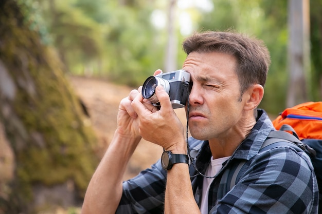 Homem focado fotografando a paisagem e andando na floresta. Viajante caucasiano explorando a natureza, segurando a câmera, tirando foto e carregando a mochila. Conceito de turismo, aventura e férias de verão