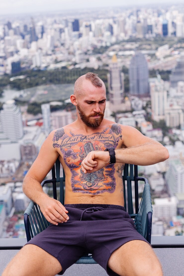 Foto grátis homem forte tatuado de barbudo europeu bonito bem sucedido em topless com relógio sentado em uma cadeira em andar alto com vista incrível da cidade