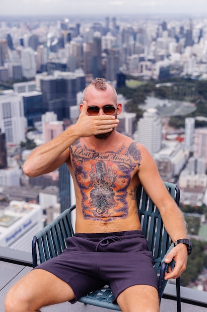 Homem forte tatuado de barbudo europeu bonito bem sucedido em topless com relógio sentado em uma cadeira em andar alto com vista incrível da cidade