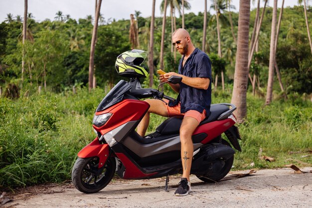 Homem forte tatuado em campo de selva tropical com moto vermelha