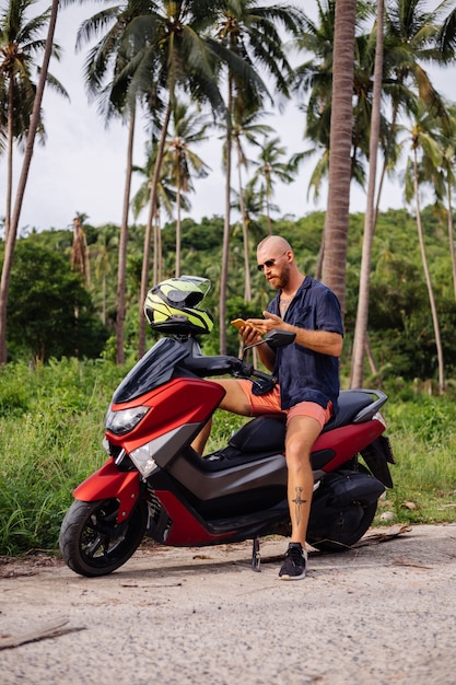 Homem forte tatuado em campo de selva tropical com moto vermelha