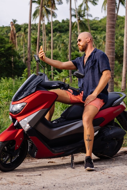 Homem forte tatuado em campo de selva tropical com moto vermelha