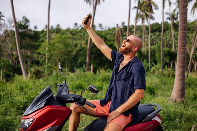 Homem forte tatuado em campo de selva tropical com moto vermelha