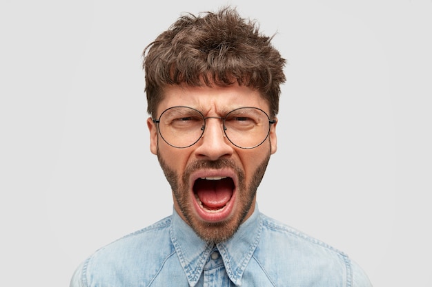 Homem furioso grita furiosamente, mantém a boca bem aberta, sente uma dor terrível, vestido com uma camisa jeans