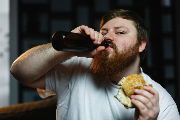 Homem gordo assiste TV, come hambúrguer e bebe cerveja