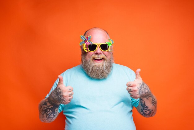 Homem gordo e feliz com tatuagens de barba e óculos de sol está pronto para o verão