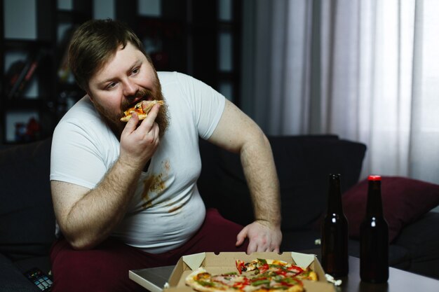 Homem gordo feio come pizza sentado no sofá