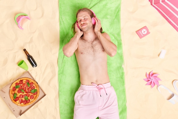 Homem gosta de ouvir suas músicas favoritas com fones de ouvido enquanto está deitado em uma toalha verde na praia, cercado por vários itens, tem um bom descanso durante as férias de verão
