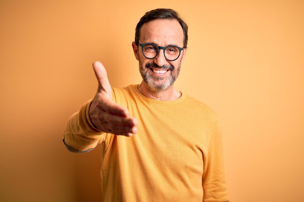 Homem grisalho de meia-idade vestindo suéter casual e óculos sobre fundo amarelo isolado sorrindo amigável oferecendo aperto de mão como saudação e boas-vindas negócio bem-sucedido