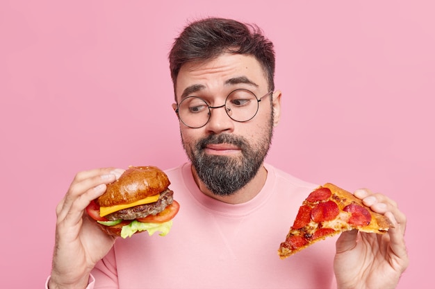 homem hesita em comer hambúrguer ou pizza prefere comer junk food usa óculos redondos e macacão