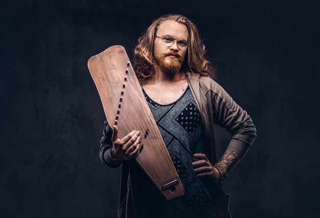 Homem hipster ruivo com cabelos longos e luxuriantes e barba cheia, vestido com roupas casuais, segura um instrumento musical tradicional russo - gusli. Isolado em um fundo escuro.