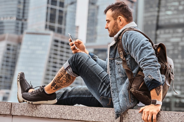 Homem hipster tatuado vestido na moda com uma mochila usando smartphone sentado na frente de arranha-céus na cidade de Moscou na manhã nublada.