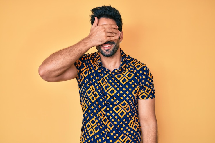 Foto grátis homem hispânico bonito com barba vestindo camisa estilo discoteca dos anos 80 sorrindo e rindo com a mão no rosto cobrindo os olhos para o conceito cego de surpresa