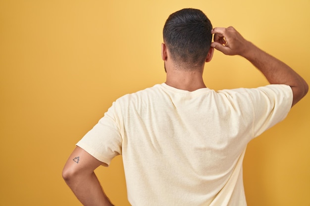 Foto grátis homem hispânico bonito em pé sobre fundo amarelo para trás pensando em dúvida com a mão na cabeça