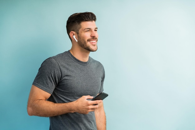 Homem hispânico bonito olhando para longe e segurando o celular enquanto ouve música através de fones de ouvido contra fundo liso