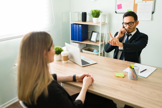 Homem hispânico bonito usando óculos e um terno falando ao telefone com um cliente enquanto diz a uma colega de trabalho caucasiana para esperar um minuto