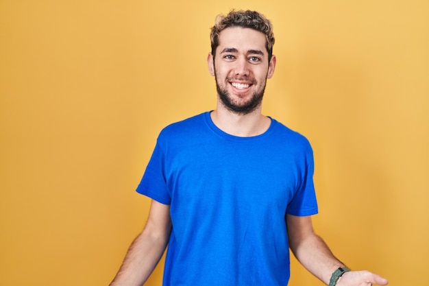 Homem hispânico com barba em pé sobre fundo amarelo sorrindo alegre de braços abertos como saudações positivas e confiantes de boas-vindas amigáveis