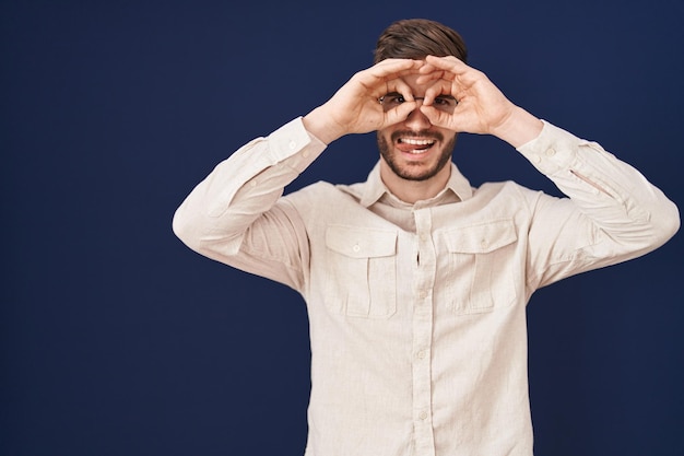 Homem hispânico com barba em pé sobre fundo azul fazendo gesto ok como binóculos enfiando a língua para fora olhos olhando por entre os dedos expressão louca