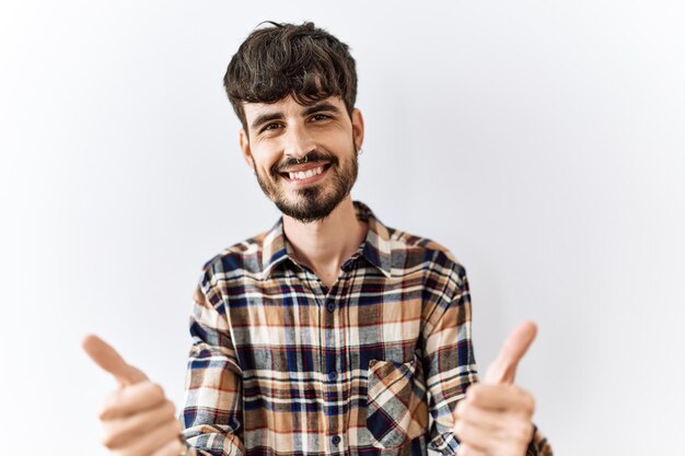 Homem hispânico com barba em pé sobre fundo isolado aprovando fazendo gesto positivo com mão polegares para cima sorrindo e feliz pelo gesto de vencedor de sucesso