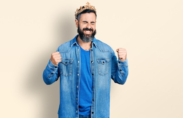 Foto grátis homem hispânico com barba usando coroa rei muito feliz e animado fazendo gesto vencedor com os braços levantados, sorrindo e gritando para o sucesso. conceito de celebração.