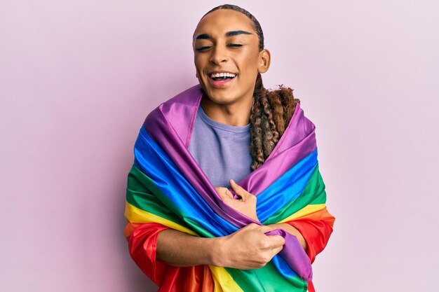 Homem hispânico usando maquiagem e cabelos longos enrolados na bandeira lgbtq do arco-íris, sorrindo e rindo muito alto porque uma piada maluca e engraçada.