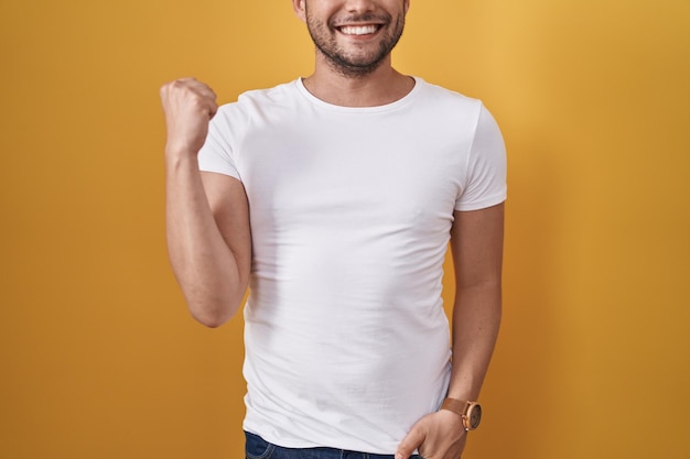 Homem hispânico vestindo camiseta branca sobre fundo amarelo gritando orgulhoso, comemorando a vitória e o sucesso muito animado com os braços erguidos