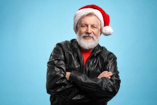 Homem idoso de cabelos grisalhos com barba real e expressão facial positiva, vestido com chapéu de Papai Noel vermelho e jaqueta de couro preta, mantendo as mãos cruzadas. Último homem posando em estúdio com fundo azul
