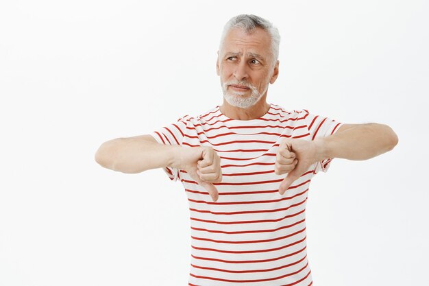 Homem idoso decepcionado com uma camiseta mostrando o polegar para baixo e mostrando gesto de desagrado