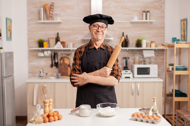 Homem idoso vestindo chef bonete sorrindo na cozinha de casa. Padeiro aposentado em uniforme de cozinha preparando ingredientes de pastelaria na mesa de madeira, pronto para cozinhar massas, bolos e pães saborosos caseiros