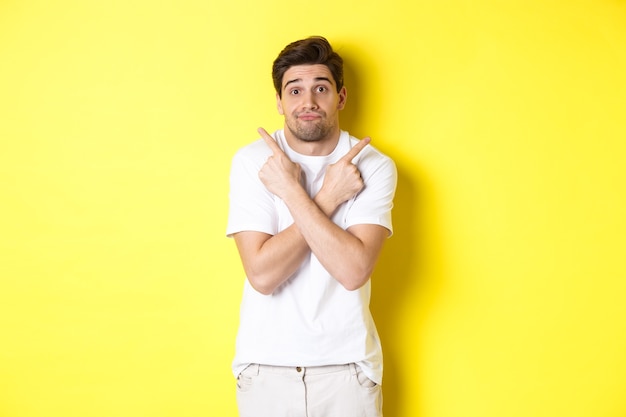 Homem indeciso apontando os dedos para o lado, lutando para tomar decisões, pedindo conselhos, de pé sobre um fundo amarelo.