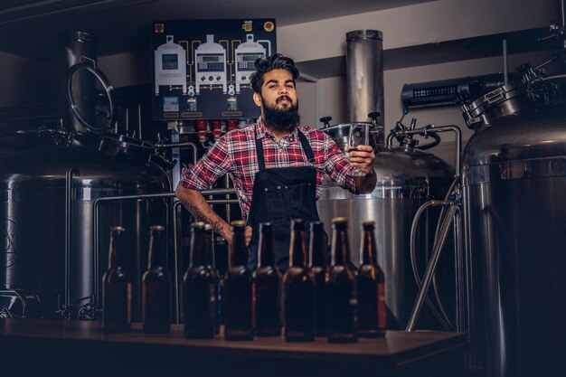 Homem indiano barbudo elegante em uma camisa de lã e avental segura um copo de cerveja, atrás do balcão em uma cervejaria.