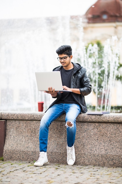 Homem indiano bonito com laptop enquanto está sentado perto da fonte no centro da cidade em um dia