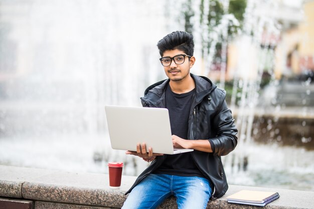 Homem indiano bonito com laptop enquanto está sentado perto da fonte no centro da cidade em um dia