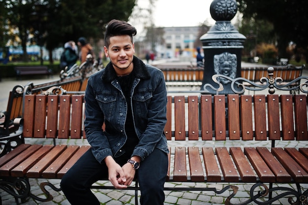 Homem indiano bonito e elegante na jaqueta jeans preta posou ao ar livre sentado no banco