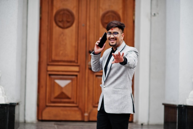 Homem indiano elegante com bindi na testa e óculos usam terno cinza posou ao ar livre contra a porta do prédio falando no celular e mostrar a mão na câmera