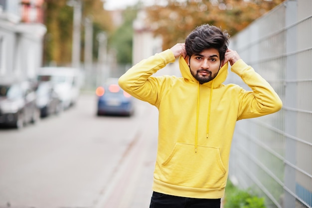 Homem índio jovem hipster urbano em um moletom amarelo na moda Cara sul-asiático legal usa capuz andando na rua de outono