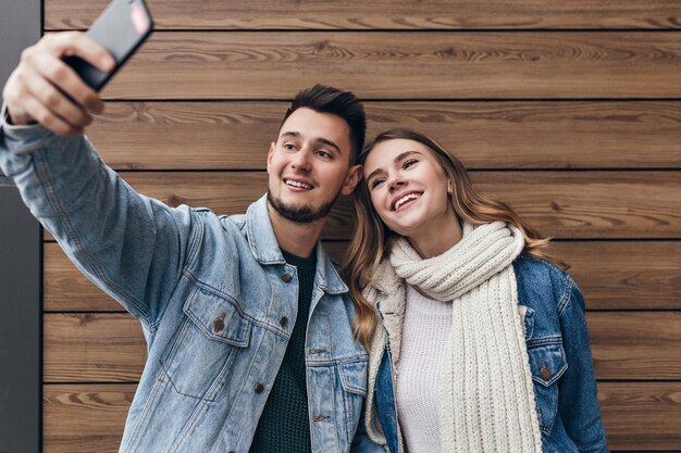 Homem inspirado com barba fazendo selfie com a namorada. Linda jovem com lenço preto, posando na parede de madeira.
