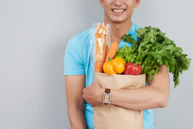 Homem irreconhecível, posando com saco de papel cheio de legumes frescos, ervas verdes e baguete
